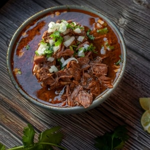 Barbacoa servida en un recipiente de vidrio. Se ven los trozos de carne, el jugo de la cocción, cebolla cruda y cilantro. Sobre la mesa de madera también hay limón verde.