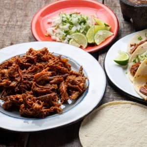 Barbacoa servida en un plato blanco. Alrededor también hay tortillas de trigo, un plato rojo con cebolla cruda en cubos y limón verde, y tacos armados con todos esos ingredientes.
