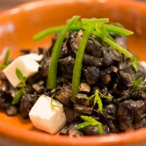 Huitlacoche guisado servido con cubos de queso de cabra, jalapeño en tiras y hojas de epazote en un plato sopero anaranjado.