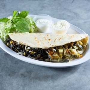 Quesadilla de huitlacoche y otras verduras guisadas, servida con tortilla de trigo junto a una ensalada de lechuga y epazote y con potes de crema ácida y queso para agregarle a gusto. El plato blanco está apoyado sobre una mesa gris.