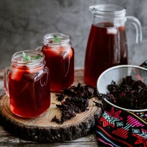 Agua fresca de jamaica servida con hielo y menta fresca en vasos de vidrio tipo frascos, con borde de rosca y asa. Están apoyados sobre una tabla, acompañados por flores de jamaica y una jarra con más de esta bebida. También se ve un matel típico mexicano.