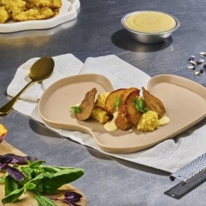 Postre servido en plato irregular color beige, apoyado sobre una servilleta blanca y con una cuchara dorada. El platillo está formado por diferentes capas de bizcocho de limón, láminas de duraznos caramelizados, ganache de albahaca y tostaditas de maíz blanco, decorado con hojas de menta fresca. Sobre la mesada de acero inoxidable se ven ingredientes extra, como hojas de albahaca fresca y un contenedor con más ganache de albahaca.