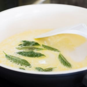 Hojas de salvia sumergidas en mantequilla derretida, calentándose en un sartén de interior blanco y exterior negro. Para remover, se utiliza una espátula blanca.