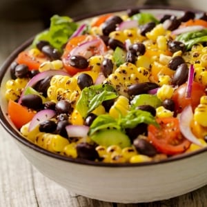 Ensalada de elote asado desgranado, frijoles negros, jitomate, cebolla morada y hojas verdes servida en un bol blanco con borde oscuro, apoyado sobre una mesa de madera. Ensalada de elote asado desgranado, frijoles negros, jitomate, cebolla morada y hojas verdes servida en un bol blanco con borde oscuro, apoyado sobre una mesa de madera.