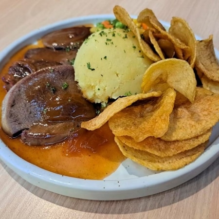 Plato servido con lengua de res en salsa, acompañado de puré de papa y chips de plátano.