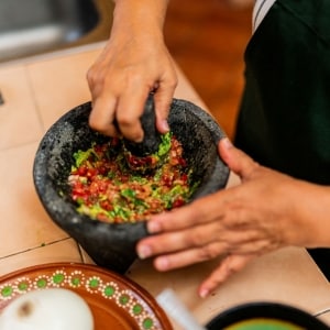 Una cocinera prepara guacamole de forma tradicional, martajando en el molcajete cebolla, ajo, chiles, jitomate y aguacate. Sobre la mesada cerámica también se ven platos con ingredientes.