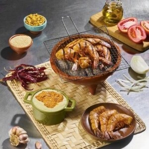 Camarones enteros asados en un grill de mesa, servidos aparte en un plato marrón junto con un puré de papa y salsa martajada en un recipiente de cerámica verde. Todos los recipientes están sobre un petate, en el que también hay chiles de árbol enteros. Alrededor, sobre la mesa de acero inoxidable, hay otros ingredientes frescos.