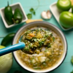 En una cuchara metálica color turquesa se muestra el resultado final de la salsa martajada, preparada con jalapeños, tomate verde, ajo, cebolla y cilantro, con una textura rústica. De fondo se ve el bol con el resto de la salsa, y alrededor hay ingredientes frescos.