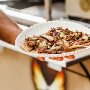 Hombre sostiene con su mano un plato blanco con cuatro tacos de carnitas, servidos con tortillas, trozos de carne de res y salsa verde.