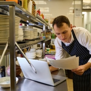 Cocinero analiza las páginas de su menú al tiempo que utiliza su computadora, que está apoyada sobre la mesa de trabajo, junto al anaquel que guarda los platos, utensilios y otra vajilla. Viste camisa blanca y mandil oscuro con rayas blancas.