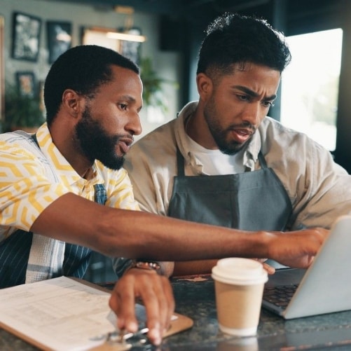 Dos hombres empleados del restaurante analizan el inventario del restaurante usando un anotador y una computadora portátil. 