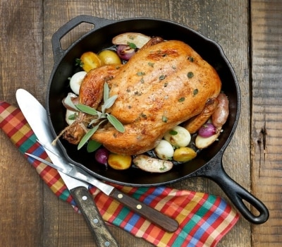 Pollo entero asado en una fuente de hierro con papas, cebollas y hierbas, apoyado sobre una mesa de madera junto a los cubiertos y una servilleta de colores.