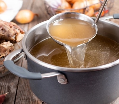 Un cucharón carga caldo de pollo de una olla gris. Alrededor se ven algunos ingredientes como huesos de pollo, cebollas y ajo.