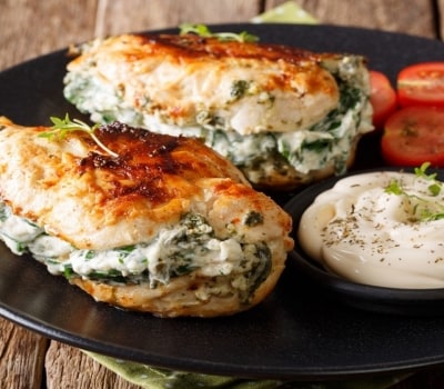 Dos pechugas de pollo doradas por fuera y rellenas de espinacas y queso, servidas junto a tomates cereza y un aderezo a base de mayonesa.