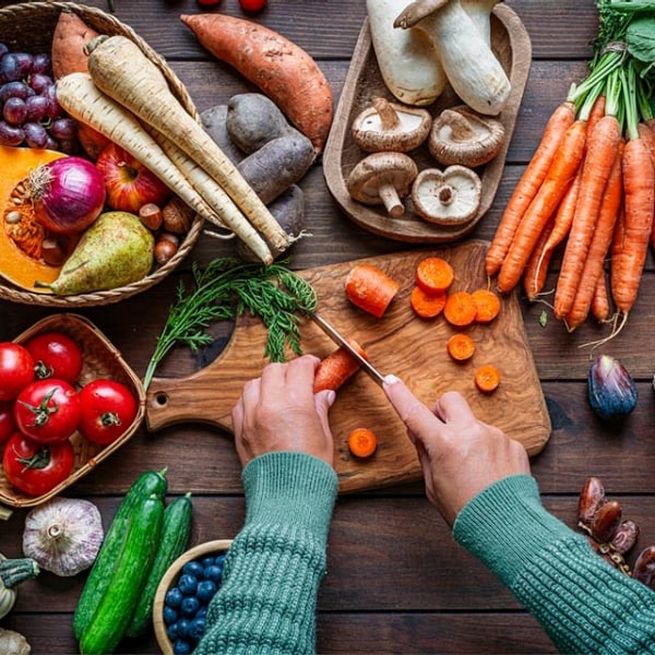 manos de mujer cocinera cortando zanahorias en rodajas con cuchillo sobre tabla de madera, alrededor de la mesa hay ingredientes varios en canastas