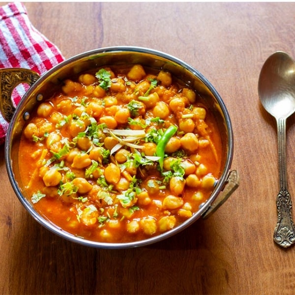 plato hondo de curry con garbanzos, tomates, cebollas y especias aromáticas con cuchara al costado sobre mesa de madera, garbanzo precocido