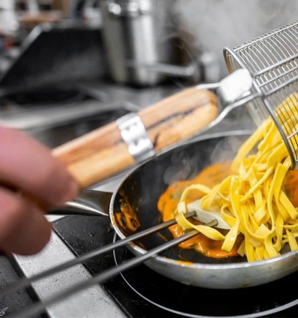 mano de cocinero volcando pasta precocida del espumadero a la sartén con salsa de tomate