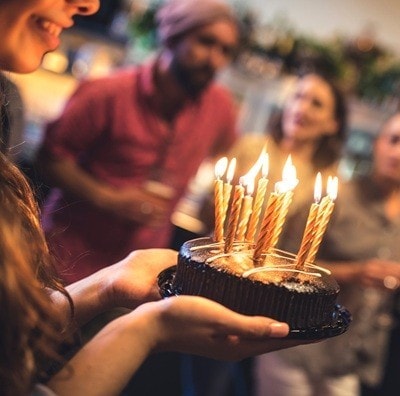 Young woman holding a birthday cake in a bar. She is ready to blow the candles. Her friends standing in back, at bar counter, and they are defocused.