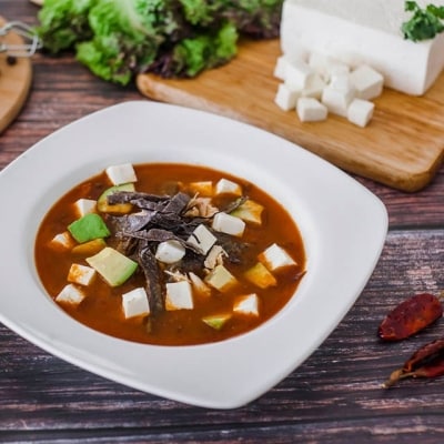 plato de sopa de tortilla mexicana con chile y queso panela en trozos junto con otros ingredientes como ají, ajo, pimienta, lechuga y queso en tablas de madera