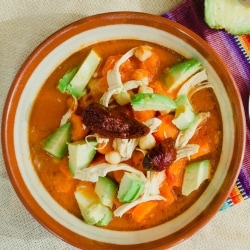 Caldo Tlalpeño servido en un plato de cerámica color ladrillo y blanco, apoyado sobre un mantel tradicional. En el consomé se ve pollo desmenuzado, zanahoria y calabaza, y está terminado con tajadas de aguacate y chipotle por encima. Caldo Tlalpeño servido en un plato de cerámica color ladrillo y blanco, apoyado sobre un mantel tradicional. En el consomé se ve pollo desmenuzado, zanahoria y calabaza, y está terminado con tajadas de aguacate y chipotle por encima.