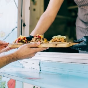 Un cocinero que usa guantes de nitrilo sirve una tabla con tres tacos diferentes a un cliente, pasando el pedido por la ventana de su food truck color azul celeste.