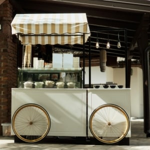 Remolque de comidas con ruedas de bicicleta instalado en un evento gastronómico, listo para ofrecer helados y otros postres. El móvil es blanco, tiene exhibidores para los productos y un toldo a rayas.