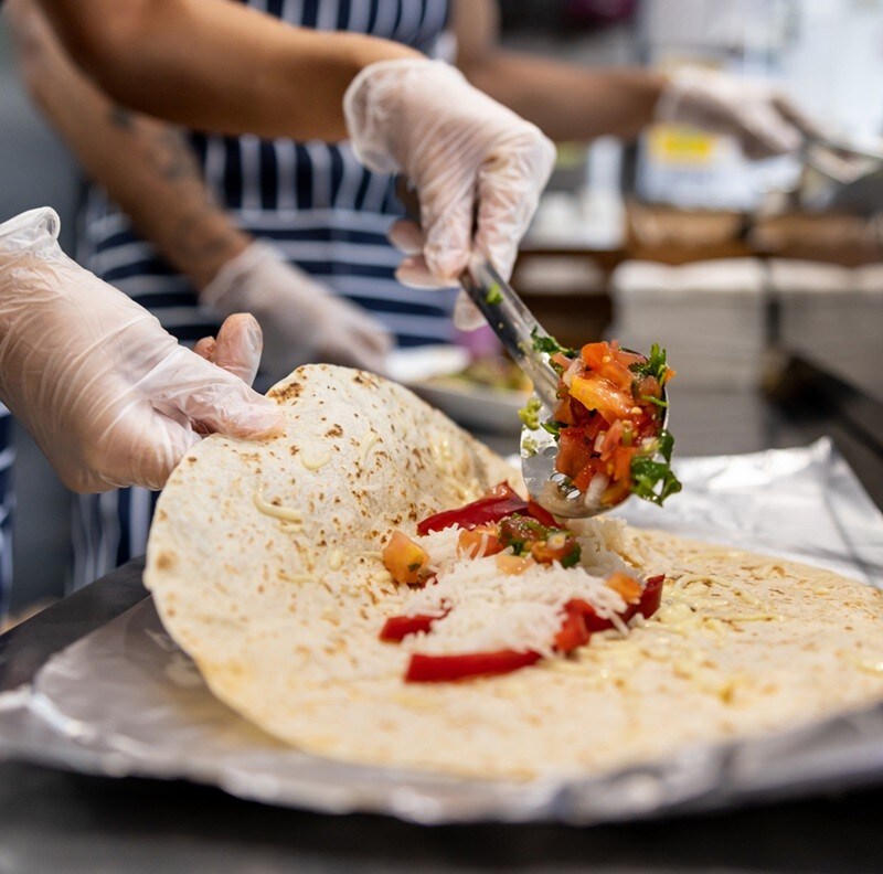Manos y brazos de personas preparando deliciosos burritos sobre una encimera. Están usando guantes, delantal y todo lo necesario para mantener la higiene durante la preparación. Manos y brazos de personas preparando deliciosos burritos sobre una encimera. Están usando guantes, delantal y todo lo necesario para mantener la higiene durante la preparación.