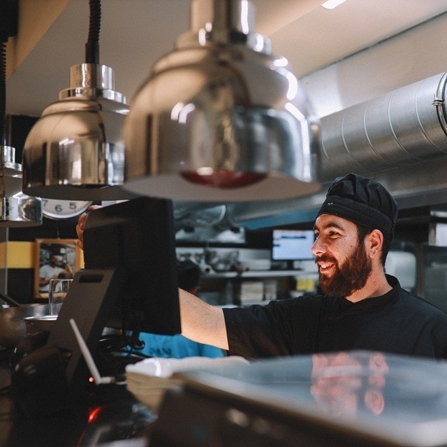 Un cocinero que viste uniforme negro sonríe mientras lee los pedidos recibidos en la pantalla del sistema POS de su cocina profesional.