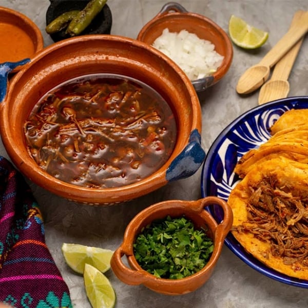  tacos de birria y caldo, perejil y chiles sobre mesada gris  