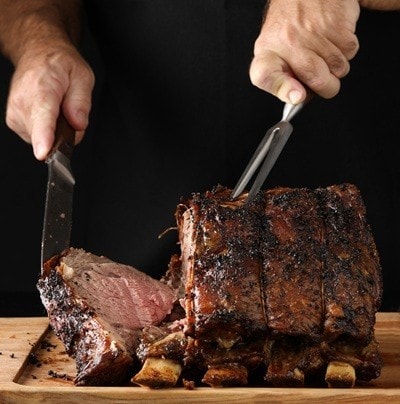 A close up vertical photograph of a chef slicing off the end piece of a freshly cooked prime rib. Isolated in black.