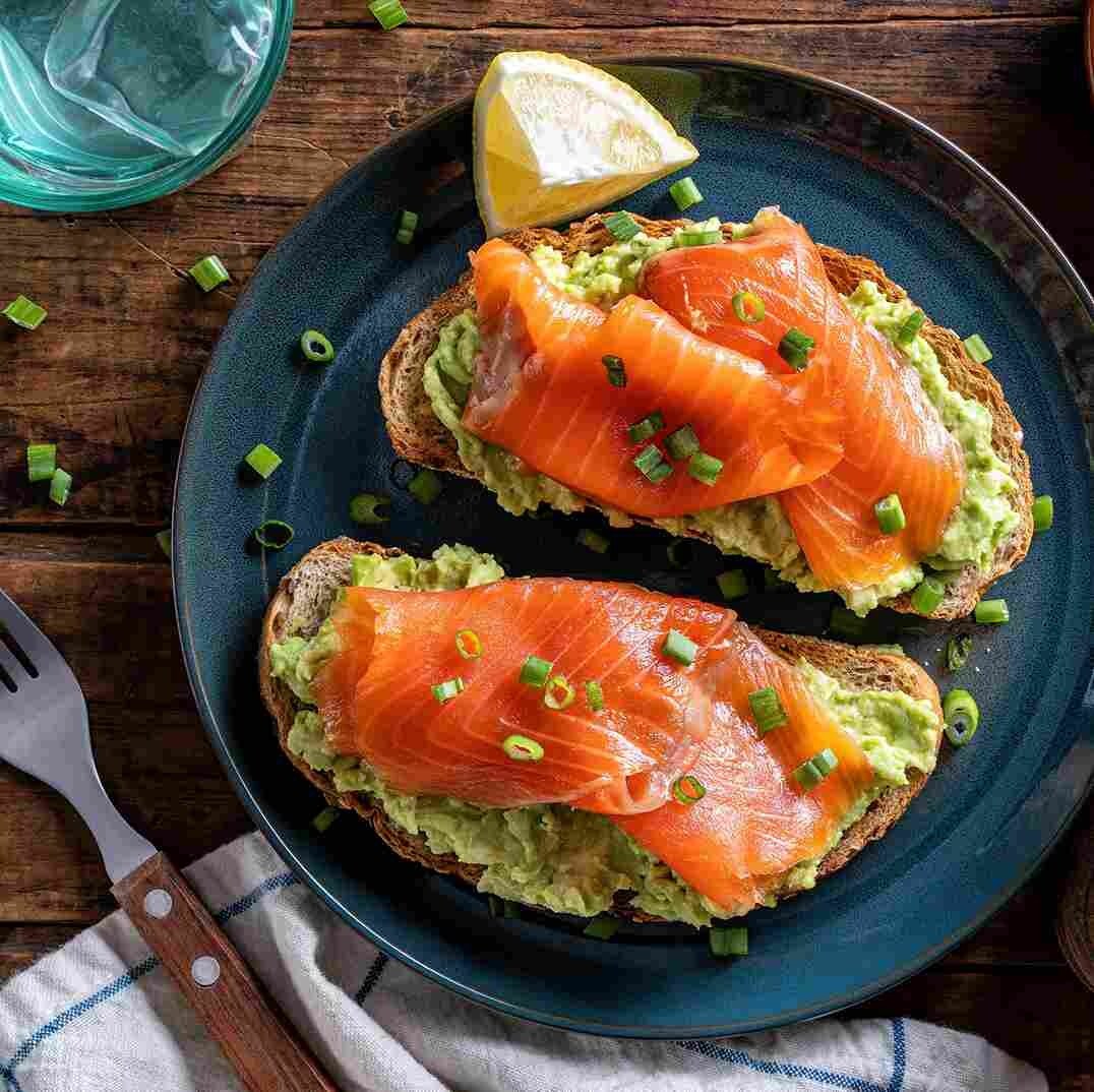 Plato con dos tostadas con salmón ahumado, aguacate y cebollín picado, servidos junto a un trozo de limón amarillo. Plato con dos tostadas con salmón ahumado, aguacate y cebollín picado, servidos junto a un trozo de limón amarillo.