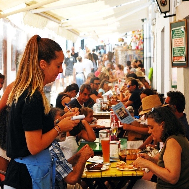 Una mesera anota un pedido en una libreta al atender las mesas llenas de comensales en la terraza exterior de un concurrido restaurante.