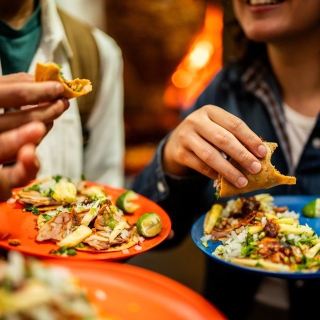 Amigos comen tacos de pie de una cantina, sosteniendo sus platos con la otra mano. 