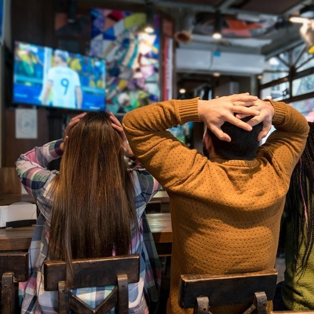 Un grupo de amigos mira fútbol en un bar, sentados en banquetas y de frente a la televisión. Se toman la cabeza reaccionando a la jugada.