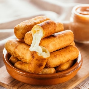 Plato de arcilla con una torre de tequeños dorados por fuera y con queso fundido por dentro. De fondo se ve un frasco de salsa rosa.