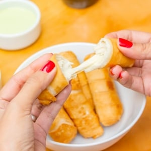 Una mujer abre a la mitad un tequeño de queso fundido usando sus dos manos con uñas pintadas de rojo. Sobre el fondo amarillo se ven un plato con más tequeños Y un contenedor con crema de guacamole para tequeños.