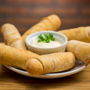 Tequeños servidos alrededor de un contenedor de aderezo a base de mayonesa y con hojas de perejil arriba. Están sobre un plato blanco y una mesa de madera.