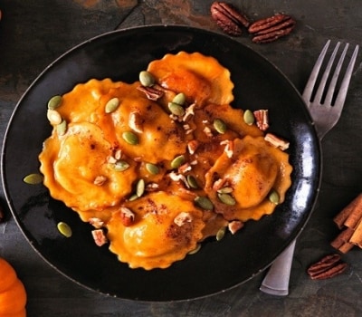 Pasta rellena de forma redonda y color naranja, con bordes ondulados, preparada con calabaza. Está servida en un plato negro con nueces partidas y semillas de calabaza.