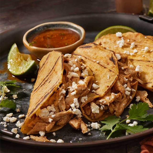 tacos de pollo con tortillas de maíz, cilantro y queso cobija sobre plato de cerámica negro con rodaja de lima y bol con salsa de tomate.  
